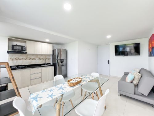 Cocina abierta y zona de comedor modernos en Olamar Living, Coveñas, con mesa de comedor, cocina equipada, sofá gris y televisor.