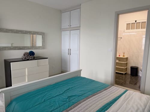 Quarto moderno no Check Madeira em Ribeira Brava com cama em tons turquesa, cômoda branca, espelho e banheiro privativo