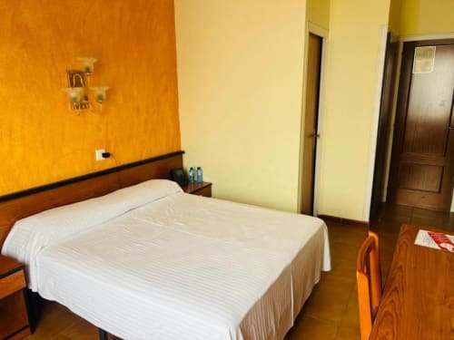 Habitación doble del Hotel Continental Calella con cama blanca, pared acentuada naranja, paredes amarillas, muebles de madera y vistas a la ciudad