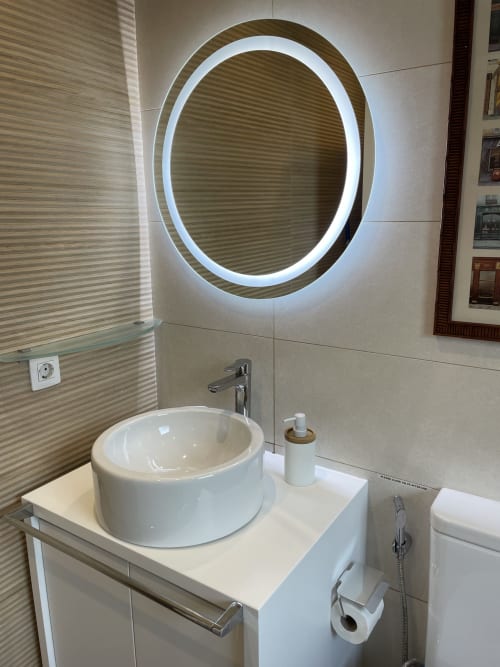 Baño moderno con espejo ovalado iluminado LED, lavamanos blanco redondo y paredes con azulejos texturados en villa de Lanzarote