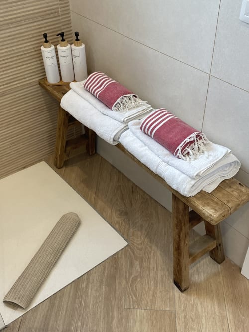 Estante de baño de madera con toallas blancas dobladas, toallas a rayas burdeos y tres botellas de artículos de tocador en baño de villa.