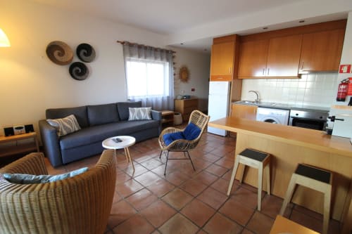 Sala de estar integrada da Villa Alegria com sofá escuro, cadeiras de vime, cozinha com armários de madeira e piso em terracota