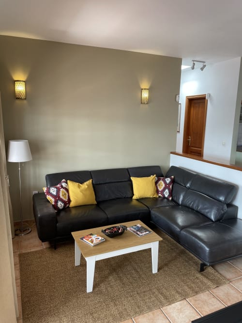 Sala moderna con sofá seccional de cuero negro, almohadas amarillas, mesita de madera y paredes beige con apliques de pared