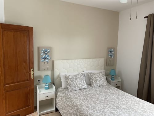 Dormitorio moderno con cabecera acolchada blanca, ropa de cama estampada, mesitas con lámparas azules, puerta de madera y arte floral en la pared