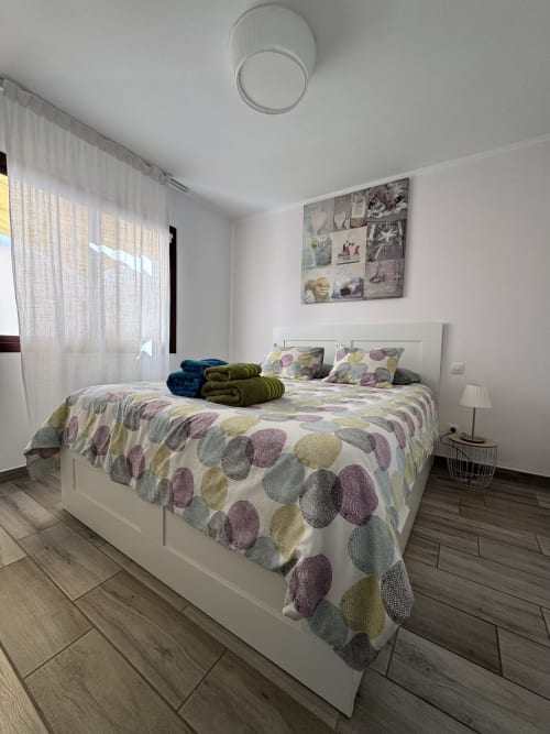 Moderno dormitorio de apartamento en Corralejo con cama blanca, ropa de cama colorida, suelos de madera y luz natural de la ventana