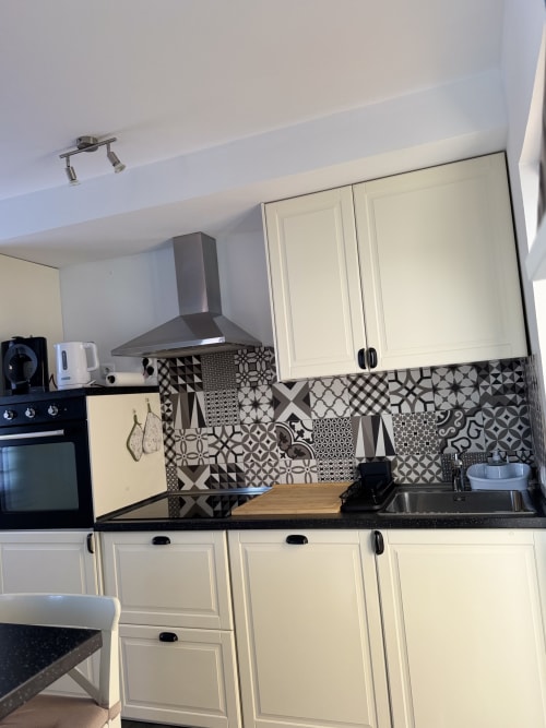 Cocina moderna con gabinetes color crema, azulejos traseros geométricos en blanco y negro, encimera oscura y campana extractora de acero inoxidable