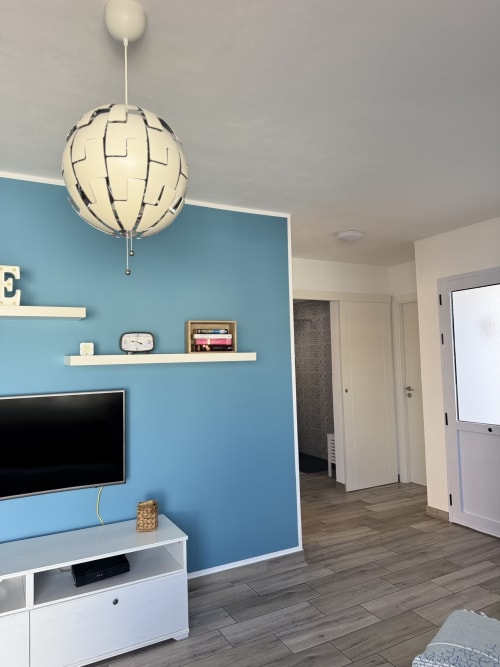 Moderno salón con pared de acento azul, televisor, lámpara colgante y estantería blanca en Corralejo, ES.