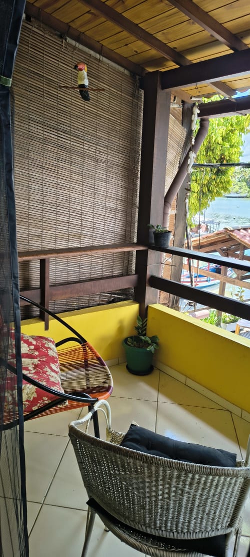 Área de estar na varanda da Casa do Porto com cadeira em vime, paredes amarelas e vista para o mar em Boipeba, Brasil