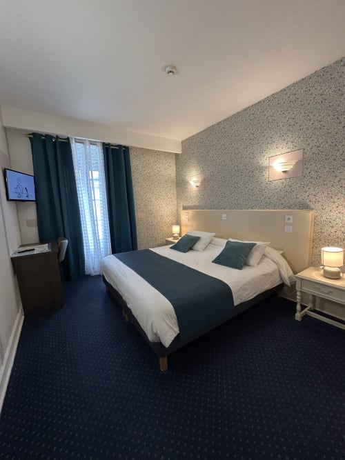 Modern hotel bedroom at Le Centre in Les Herbiers with queen bed, dark carpet, patterned wallpaper, and city views from