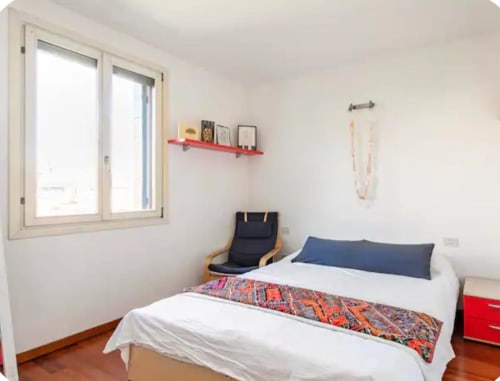 Camera luminosa presso Mappamondi B&B a Venezia con letto bianco, biancheria colorata, scaffale rosso, pavimento in legno e ampia finestra.
