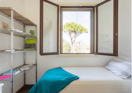 Camera luminosa al Mappamondi B&B di Venezia con finestra aperta sulla vegetazione, letto bianco, coperta blu e scaffali metallici