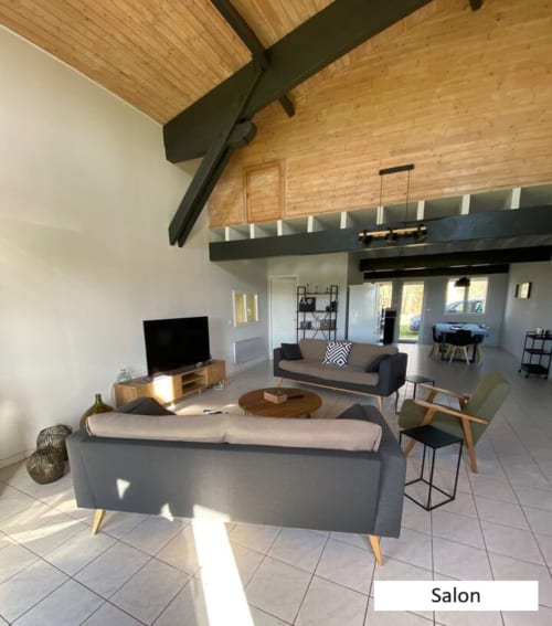 Salon contemporain du gîte De Bigorre à Mauvilla avec canapés gris, poutres en bois et vue sur jardin à Civrac sur Dordogne