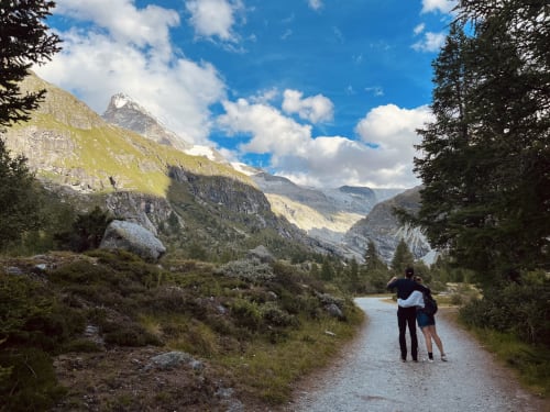 Deux randonneurs s'enlacent sur un sentier montagnard près de La Sage, avec des pics enneigés et des alpages en arrière-plan