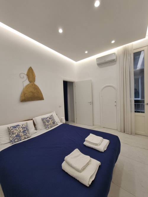 Camera suite moderna con letto blu navy, cuscini bianchi, arte murale dorata, illuminazione incassata, unità AC e porta in vetro verso il balcone