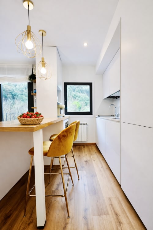 Cocina moderna en apartamento Holibai Lisiña, Baiona con muebles blancos, lámparas colgantes doradas y vistas a la montaña