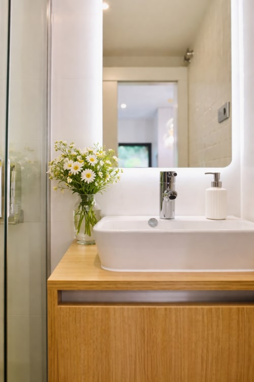 Baño moderno en apartamento Holibai, Baiona con lavabo blanco, mueble de madera, flores de margarita y espejo iluminado