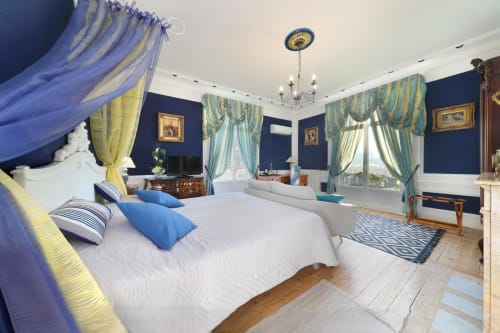 Chambre luxueuse avec lit à baldaquin bleu, tentures jaunes et turquoise, murs bleu marine et vue sur jardin au Château de Quesmy