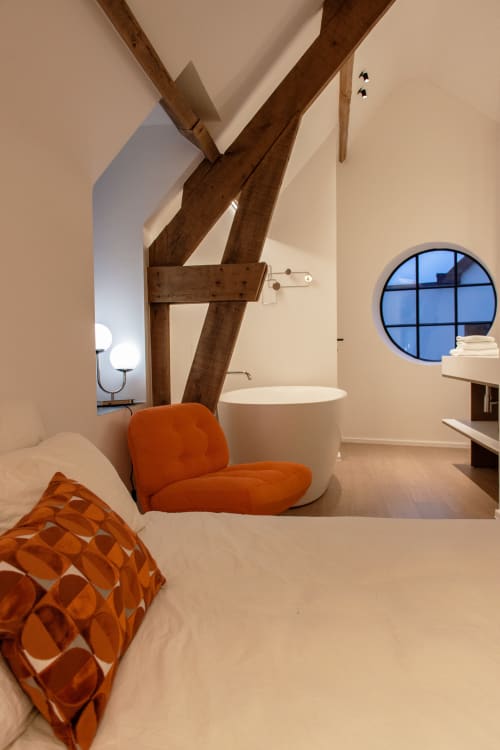 Modern bedroom at Castanea Business & Suites with exposed wooden beams, orange accent chair, and circular window in Merelbeke