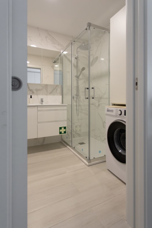 Baño moderno con ducha a ras de suelo, paredes de mármol, tocador blanco y lavadora integrada en apartamento de Sesimbra.
