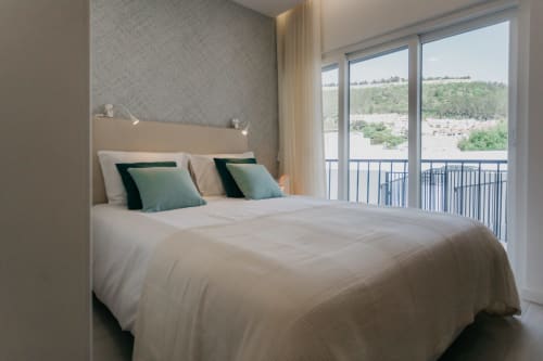 Dormitorio moderno en apartamento de Sesimbra con vista al río y ladera verde, ropa de cama color crema, almohadas verde azulado, acceso a balcón