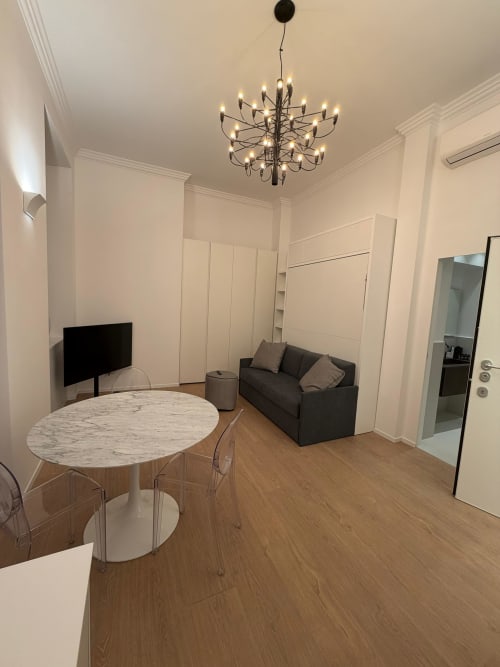 Modern studio apartment at Maisons Milano featuring marble dining table, dark sofa, TV, and contemporary chandelier
