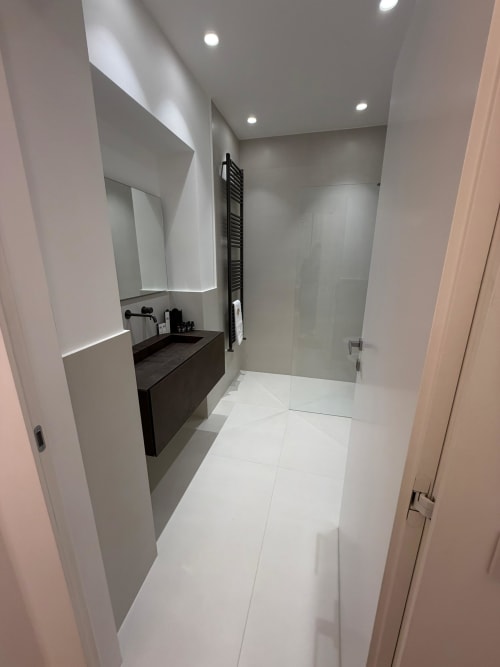 Modern bathroom at Maisons Milano with dark vanity, illuminated mirror, white tile flooring, and glass shower enclosure