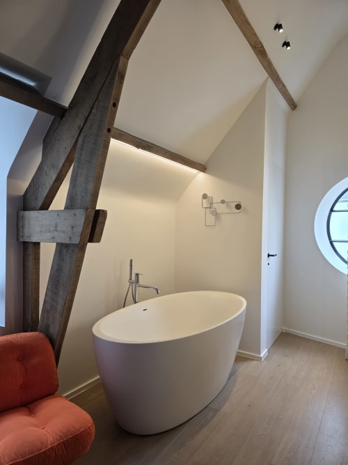 Modern bathroom with freestanding white bathtub, exposed wooden beams, and circular window at Castanea Studio Fabienne,