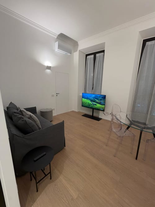 Soggiorno di appartamento moderno a Milano con divano scuro, televisore schermo piatto, sedie trasparenti, pavimenti in legno e arredamento contemporaneo