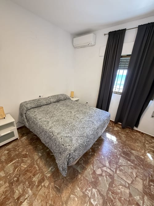 Dormitorio con cama estampada, mesita blanca, cortinas negras y suelo de mármol en apartamento frente al mar de Matalascañas