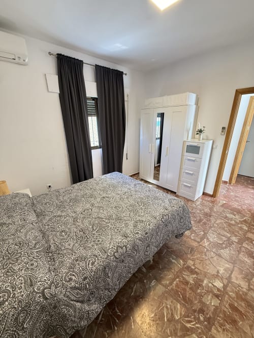 Dormitorio espacioso con colcha estampada, armario blanco, cortinas oscuras y suelo de terracota en apartamento de Matalascañas