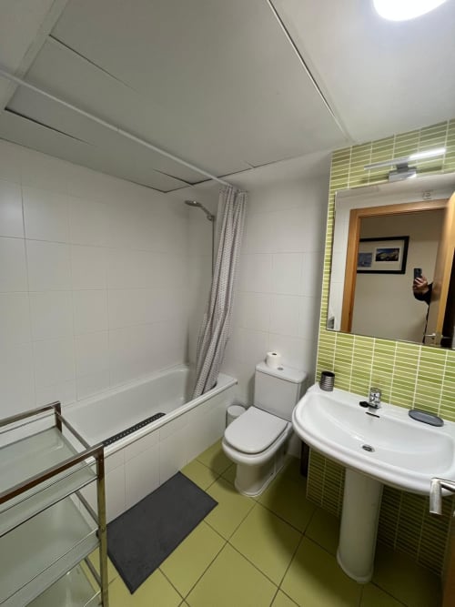 Salle de bain moderne avec accents en carrelage vert, toilettes blanches, lavabo sur colonne et baignoire avec douche à l'appartement Sant Jordi