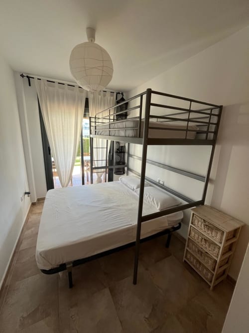 Chambre lumineuse avec lit superposé en métal, lit simple, table de nuit en osier et balcon avec vue sur le jardin dans l'appartement Sant Jordi
