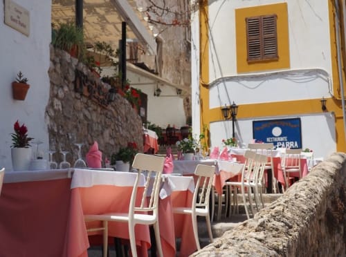Outdoor dining area at Hotel La Ventana in Ibiza with coral tablecloths, white chairs, potted flowers, and yellow