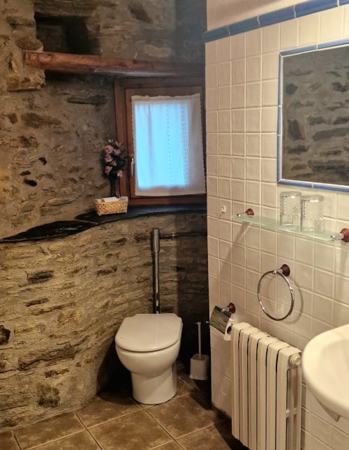 Baño rústico en Hotel Mas D'En Roqueta con paredes de piedra, inodoro blanco, radiador y pequeña ventana con cortina de encaje