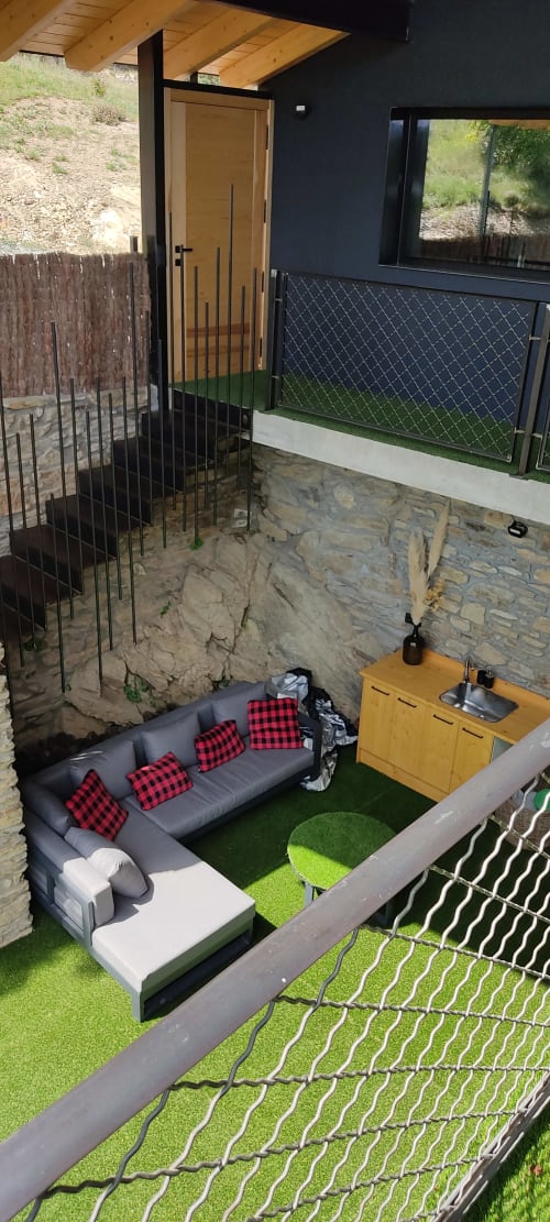 Terraza moderna con sofá gris, cojines rojos, césped artificial, kitchenette de madera, barandillas negras y vistas a montañas en Mas d'en