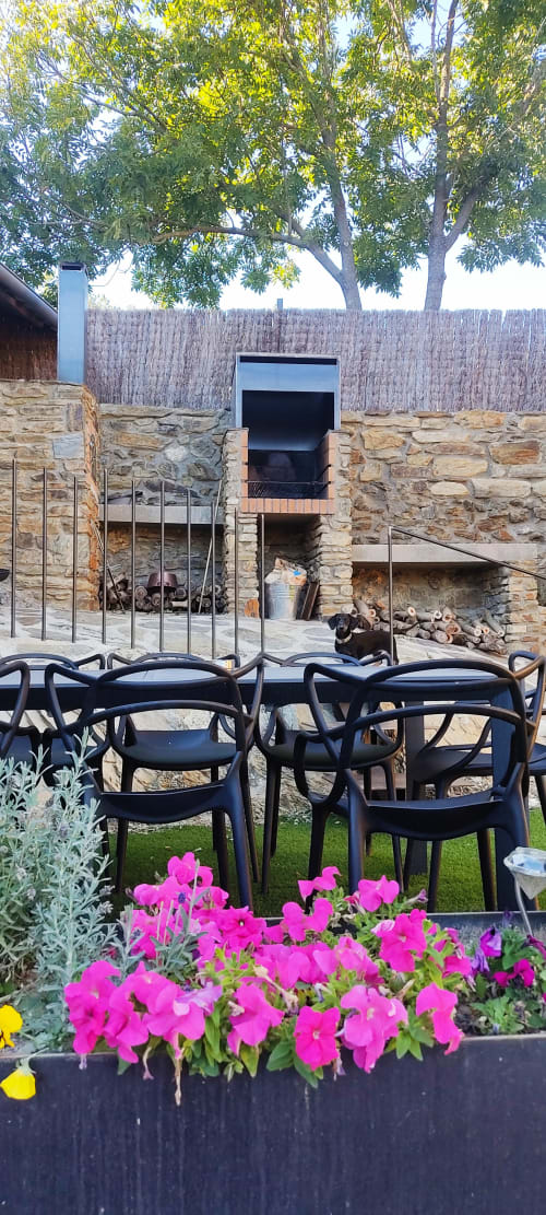 Zona de descanso exterior en la casa rural Mas d'en Roqueta con petunias rosas, sillas negras, muros de piedra y árboles frondosos