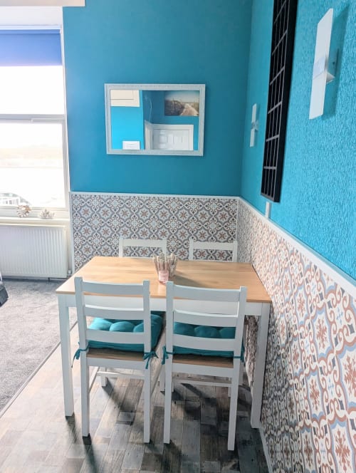 Bright turquoise dining area with wooden table, white chairs, patterned wainscoting, and sea view window in Seashell Suite at