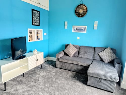 Seashell Suite sitting area with turquoise walls, gray sofa, TV, white cabinet, clock, and framed artwork at Commodore Hotel,
