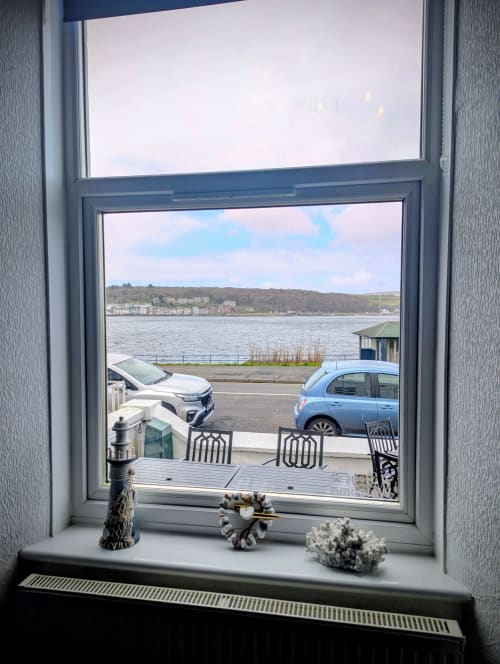 Window view from Seashell Suite showing Rothesay Bay, parked cars, waterfront, and hills across the water