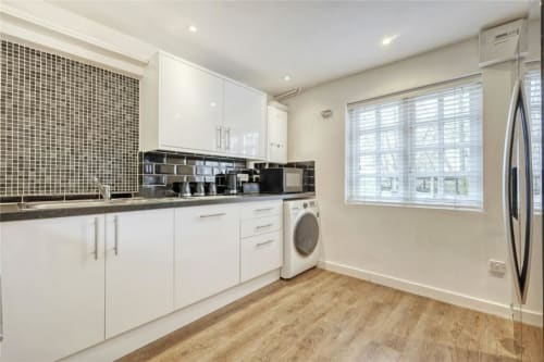 Modern white kitchen with green tile backsplash, dark countertop, washer, stainless steel refrigerator, and wooden flooring