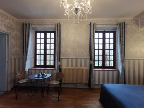 Chambre élégante au Coin des Artistes avec deux grandes fenêtres, lustre, fauteuils en rotin et literie bleue