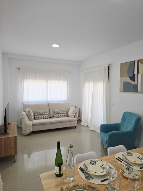 Sala de estar moderna en Gandía con sofá beis, sillón azul turquesa, comedor y amplias ventanas con vistas al jardín
