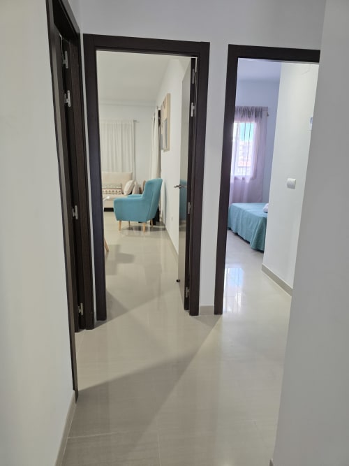 Pasillo luminoso con puertas abiertas que muestran sala con silla turquesa y dormitorio en apartamento de playa en Gandía