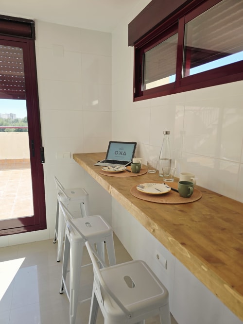 Espacio de trabajo moderno en apartamento de playa en Gandía con escritorio de madera, laptop, sillas blancas y vistas a las montañas a través de marco burdeos