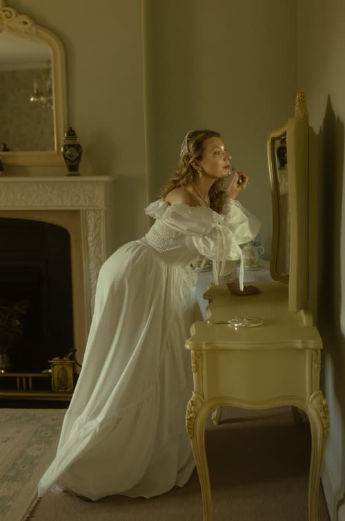 Frau im weißen Historienkleid am Frisiertisch mit Spiegel in elegantem cremefarbenem Schlafzimmer in den Florence Nightingale Suites, Derbyshire