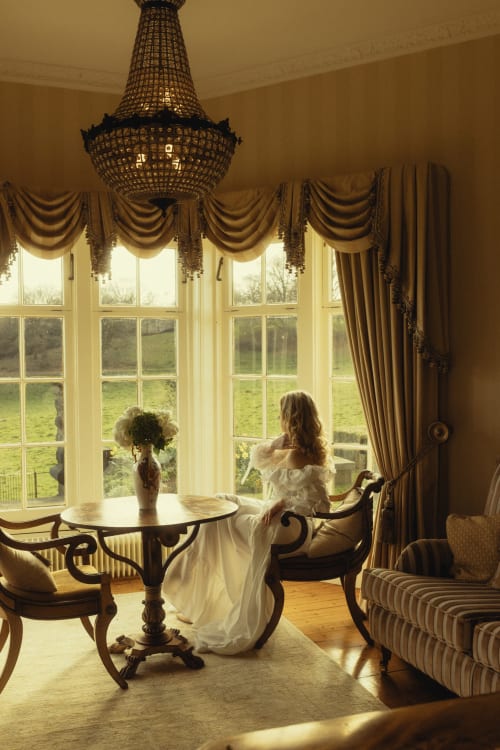 Elegante Frau im weißen Kleid blickt auf die Derbyshire-Landschaft durch große Fenster, Kristallleuchter darüber