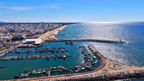 Vista aérea do hotel RB Algarve com praia de areia, águas turquesas, barcos de pesca e localidade costeira portuguesa