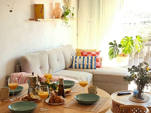 Comedor luminoso con sofá beige, mesa de madera lista para brunch, platos verdes y plantas en maceta en vivienda de Águilas.