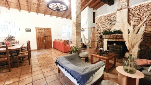 Sala de estar de la villa Bakit House en Águilas con chimenea de piedra rústica, sofá rojo, vigas de madera y suelos de terracota