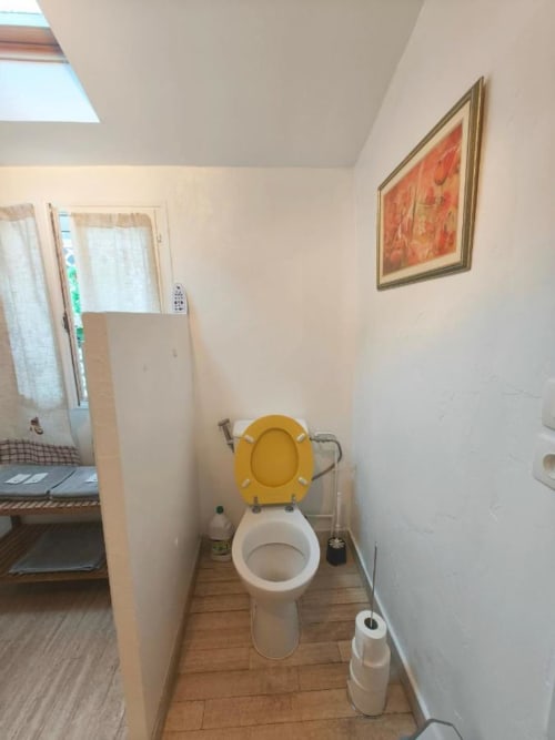 Salle de bain du Gîte Le Coin des Artistes avec siège de toilettes jaune, toilettes en porcelaine blanche, sol en bois et œuvres d'art encadrées